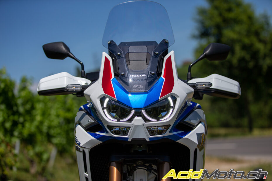 Imagem da Honda Africa Twin 1100, uma motocicleta de alto desempenho, com design robusto e características aventureiras. A moto está estacionada em um ambiente externo e é mostrada de perfil, destacando suas linhas aerodinâmicas, faróis distintivos e rodas de liga leve. O modelo exibido é na cor branca, com detalhes em AZUL e vermelho.